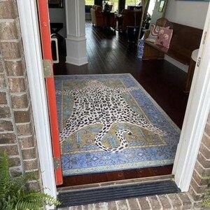 May include: A rectangular rug with a leopard print design in black and white, centered on a blue and gold patterned background. The rug is placed in a doorway, with a view of a room with dark wood floors and furniture.