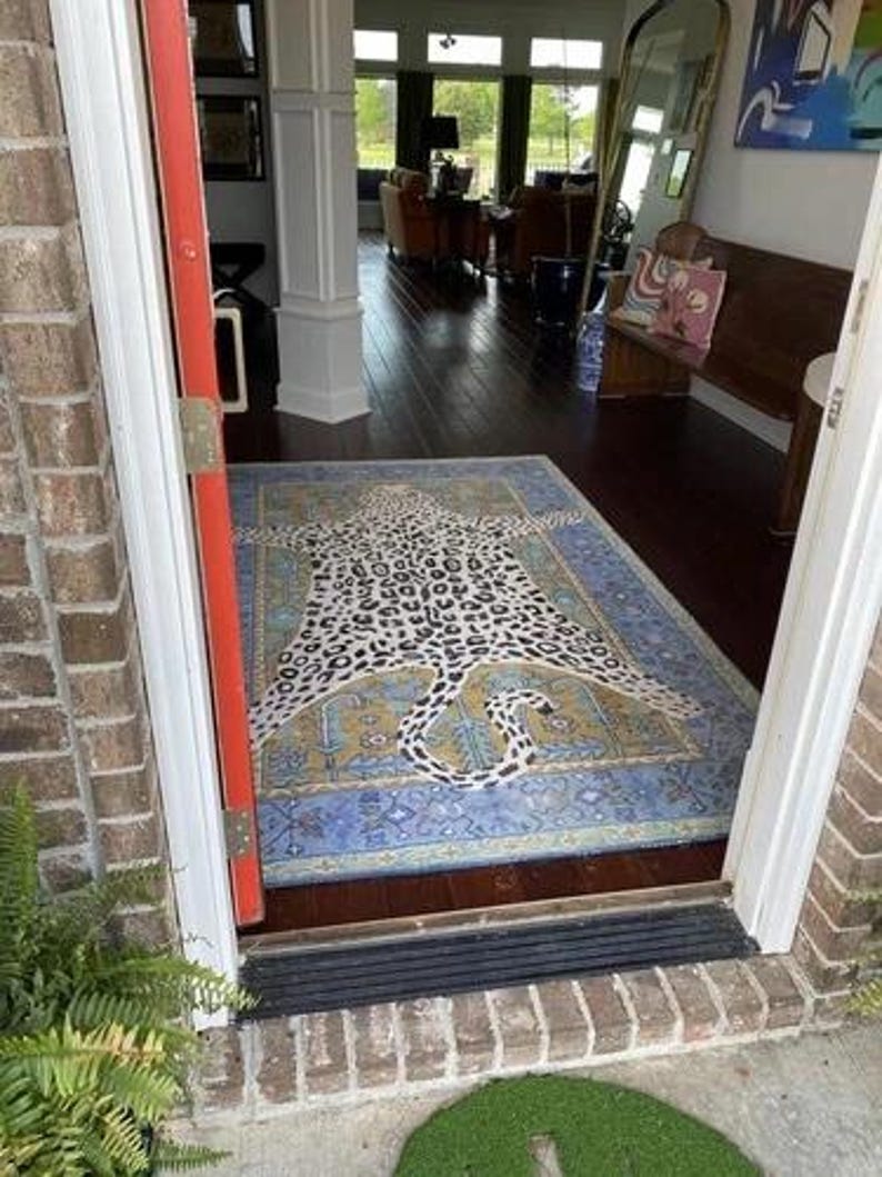 May include: A rectangular rug with a leopard print design in black and white, centered on a blue and gold patterned background. The rug is placed in a doorway, with a view of a room with dark wood floors and furniture.