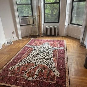 May include: A rectangular rug with a leopard print design in the center, surrounded by a burgundy border with intricate patterns. The rug is placed on a hardwood floor in a room with large windows.