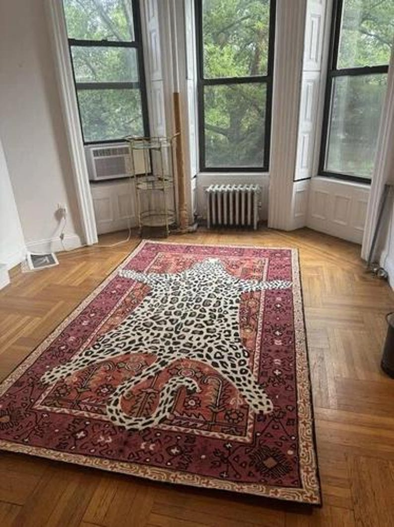 May include: A rectangular rug with a leopard print design in the center, surrounded by a burgundy border with intricate patterns. The rug is placed on a hardwood floor in a room with large windows.