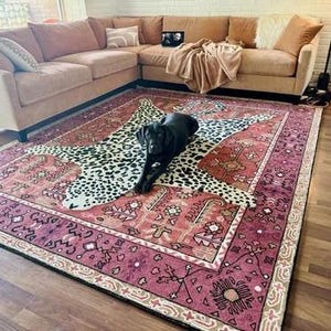 May include: A large, rectangular area rug with a detailed, patterned border in shades of burgundy, beige, and black. The rug features a central leopard print design, and a black dog is lying on top of the rug.