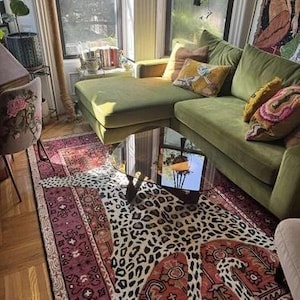 May include: A living room with a green velvet sectional sofa, a mirrored coffee table, and a leopard-print rug. The rug is layered over a patterned, burgundy-colored rug. Decorative pillows in shades of yellow and pink add color.