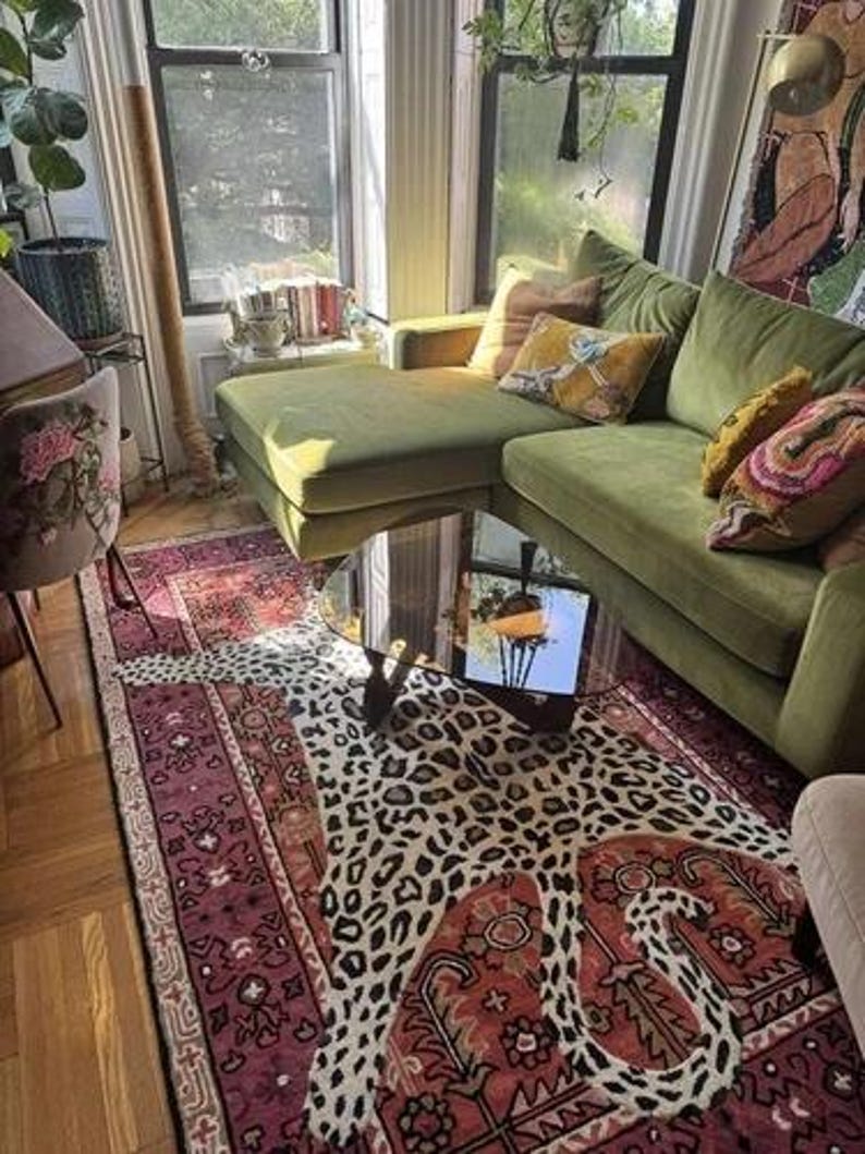 May include: A living room with a green velvet sectional sofa, a mirrored coffee table, and a leopard-print rug. The rug is layered over a patterned, burgundy-colored rug. Decorative pillows in shades of yellow and pink add color.