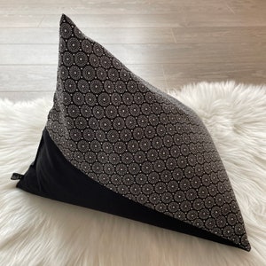 Può includere: Un cuscino decorativo con un motivo geometrico. Il cuscino presenta una base nera e una parte superiore a fantasia con cerchi e punti bianchi su sfondo nero. È appoggiato su una superficie bianca e soffice.