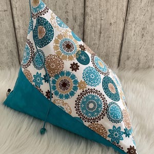 Könnte beinhalten: Ein dreieckiger Stoffbeutel mit weißem Hintergrund und einem sich wiederholenden Muster aus türkisfarbenen und beigen kreisförmigen Mandala-Motiven. Der untere Teil besteht aus einfarbigem türkisfarbenem Stoff und hat einen Kordelzug.