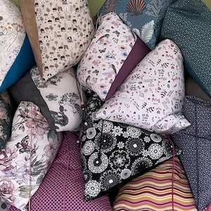 Könnte beinhalten: Eine Sammlung dekorativer Kissen mit verschiedenen Mustern und Farben, darunter florale, geometrische und abstrakte Designs. Die Kissen sind aus verschiedenen Stoffen und Texturen gefertigt, und einige haben bedruckten Text.