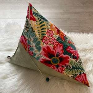 Puede incluir: Una almohada triangular con estampado floral verde y blanco con flores rojas, rosas y amarillas. La almohada tiene un cierre de cordón y una cuenta verde.