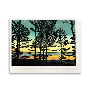 May include: A rectangular greeting card featuring a block print illustration of a forest landscape at sunset. The scene depicts black silhouettes of tall pine trees against a gradient sky of teal, yellow, and orange, with a calm body of water.