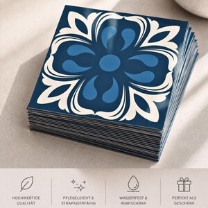 May include: A stack of square tiles featuring a dark blue background with a symmetrical, cream-colored floral pattern and lighter blue teardrop shapes. The bottom section displays four icons with the German text: HOCHWERTIGE QUALIT&Auml;T, PFLEGELEICHT & STRAPAZIERF&Auml;HIG, WASSERFEST & ABWISCHBAR, and PERFEKT ALS GESCHENK.