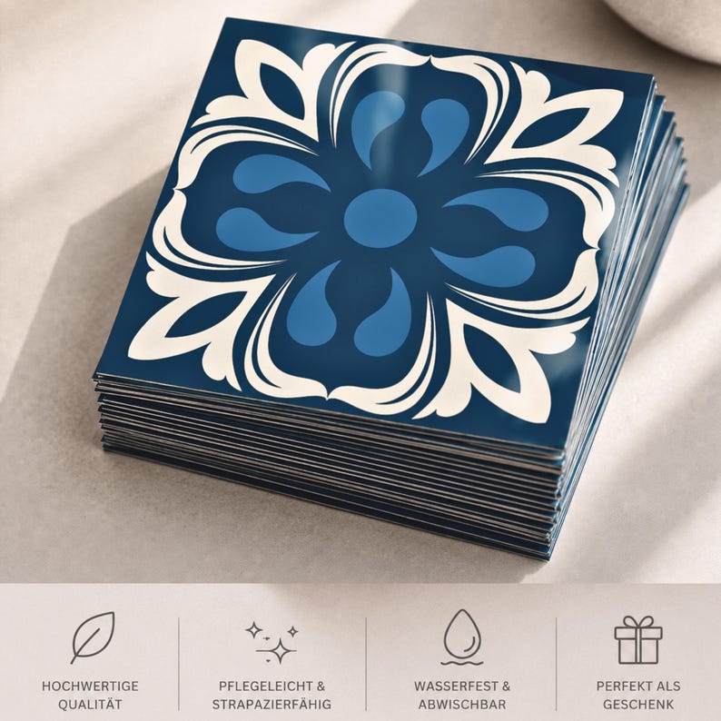 May include: A stack of square tiles featuring a dark blue background with a symmetrical, cream-colored floral pattern and lighter blue teardrop shapes. The bottom section displays four icons with the German text: HOCHWERTIGE QUALIT&Auml;T, PFLEGELEICHT & STRAPAZIERF&Auml;HIG, WASSERFEST & ABWISCHBAR, and PERFEKT ALS GESCHENK.