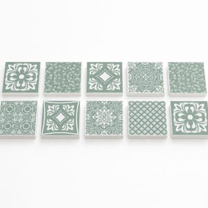 May include: A set of ten square tiles arranged in two rows of five. Each tile features a sage green background with distinct white printed patterns, including floral motifs, geometric shapes, intricate mandalas, and repeating lattice designs.
