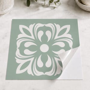 May include: A square adhesive tile decal featuring a sage green background with a centred white symmetrical floral pattern. The bottom right corner of the decal is peeled back to reveal the white adhesive backing underneath the square sheet.