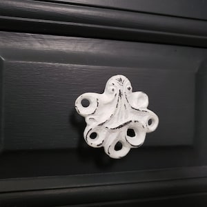 May include: White ceramic drawer pull shaped like an octopus with a distressed finish. The pull is mounted on a dark gray drawer front.