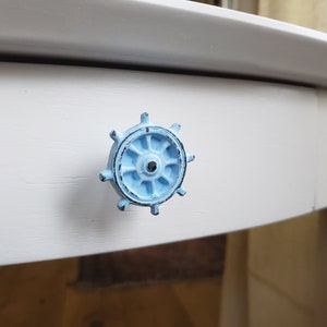 Pode incluir: Uma puxador de gaveta de estilo náutico azul claro em forma de roda de navio com um centro preto.