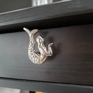 Puede incluir: Tirador de cajón de sirena plateado con un diseño detallado. La sirena está mirando hacia la derecha y tiene una cola larga y fluida.