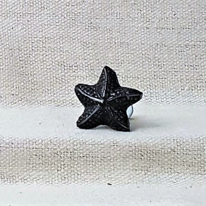 Puede incluir: Un tirador de armario negro en forma de estrella de mar. El tirador está hecho de un material oscuro y tiene una superficie texturizada. El tirador está montado sobre una superficie blanca.