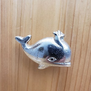 Puede incluir: Una perilla de puerta o tirador de cajón en forma de ballena plateada. La ballena está mirando hacia la derecha y tiene un ojo pequeño.
