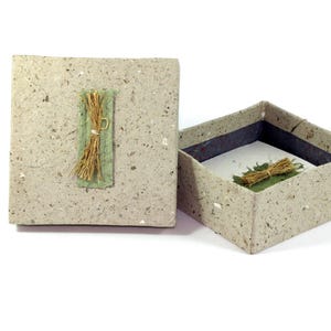 Peut inclure: Une petite boîte carrée grise avec un couvercle. La boîte est faite de papier recyclé et a un aspect naturel et rustique. Le couvercle a un détail décoratif d'un petit paquet d'herbe sèche liée avec de la ficelle.