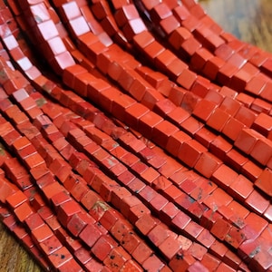Könnte beinhalten: Eine Strähne roter Jaspisperlen, jede Perle ist quadratisch mit einer leicht unregelmäßigen, rauen Textur.