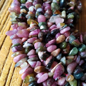 May include: A strand of multi-colored gemstone beads. The beads are small and irregular in shape, with a mix of pink, green, black, and brown hues. The beads are strung on a clear thread.