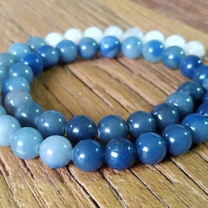 Pode incluir: Uma pulseira de contas azuis e brancas, com um gradiente de cor do claro ao escuro. As contas redondas e polidas estão dispostas em um padrão circular. A joia repousa sobre uma superfície de madeira.