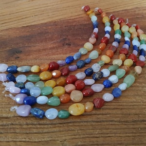 Op de afbeelding: Slingers van kleurrijke edelsteen kralen in regenboogkleuren. De ovale, gladde kralen zijn rood, oranje, geel, groen, blauw en paars. Ze zijn geregen op een transparante draad en liggen op een houten oppervlak.