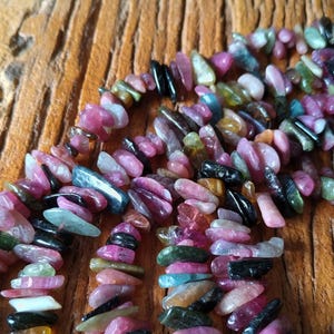 May include: A collection of tourmaline gemstone beads in various colours, including pink, green, black, and blue. The beads are strung together, forming a vibrant jewellery component. The stones are displayed on a wooden surface.