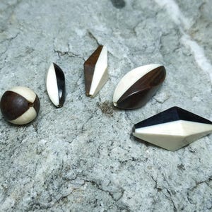 May include: A selection of handcrafted wooden beads in various shapes and sizes. The beads feature dark brown, black, and cream colours. Shapes include a sphere, teardrop, and geometric designs, showcasing a natural and artistic aesthetic.