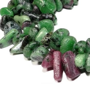 Puede incluir: Una hebra de cuentas de piedras preciosas verdes y rosas. Las cuentas tienen una forma irregular y una textura rugosa y natural.