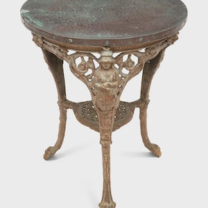 May include: Antique round side table with a textured, dark copper-colored top. The table features ornate, cast-iron legs and a decorative apron with a central figure. The table is approximately 24 inches in diameter and 28 inches tall.