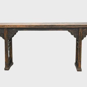 May include: A long, narrow wooden console table with a dark, distressed finish. The table features a flat top with slightly upturned ends, supported by two vertical legs connected by decorative carved brackets with intricate scrollwork patterns.