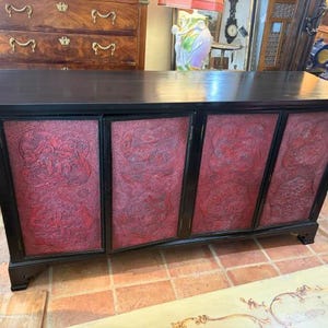 May include: A rectangular black wooden sideboard featuring four cabinet doors with inset red panels. Each red panel displays intricate, raised relief carvings of figures and landscape scenes. The sideboard has a smooth top surface and base.