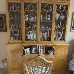May include: A large, light-colored wooden cabinet with glass doors, displaying various figurines and decorative items. The cabinet has a detailed design and is topped with a decorative edge. Below the cabinet are framed photos and a wooden chair.