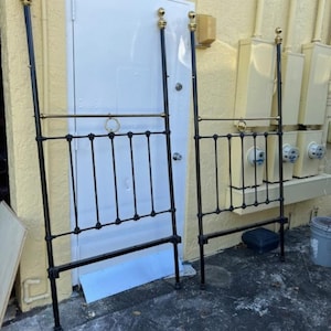 May include: Two black metal bed frames with gold accents. The frames are standing upright against a yellow wall.