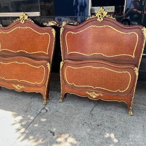 Könnte beinhalten: Zwei antike Bettrahmen aus Holz mit aufwendigen goldenen Verzierungen. Die Kopf- und Fußteile haben ein geschwungenes Design mit einer satten braunen Oberfläche. Die Rahmen weisen dekorative goldene Akzente auf.
