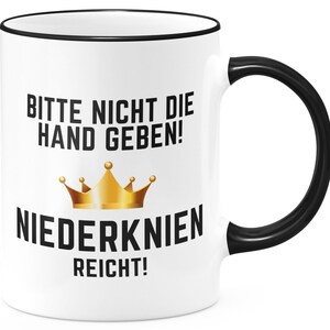 Könnte beinhalten: Weiße Keramiktasse mit schwarzem Rand und Henkel. Die Tasse hat schwarzen Text, der "Bitte nicht die Hand geben! Niederknien reicht!" lautet, mit einer goldenen Kronengrafik.