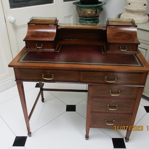 May include: A mahogany writing desk with a leather top and brass accents. The desk has two drawers on the top and four drawers on the right side. The desk is in a room with white tiled flooring.