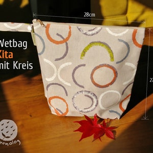 Op de afbeelding: Een beige natte tas met een ritssluiting en een kleurrijk abstract patroon van cirkels en bogen in tinten oranje, grijs en groen. De tas meet 28 cm bij 27,5 cm en heeft de tekst "Wetbag Kita mit Kreis" op de voorkant.