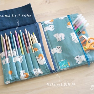 Op de afbeelding: Een blauwe en witte stoffen potlooddoos met een ritssluiting. De doos heeft een patroon van ijsberen en oranje ringen. De doos is open en gevuld met kleurpotloden, stiften en een schaar. De tekst "Maximal bis 15 Stifte" staat op de doos gedrukt. De tekst "Malblock DIN A5" staat op de doos gedrukt.