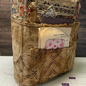 Può includere: Un diario in tessuto fatto a mano con un'estetica vintage. Il diario presenta una parte superiore a fantasia floreale, un corpo in velluto beige e dettagli in pizzo. L'interno include carta invecchiata e una tasca con inserti floreali. Il design combina trame e colori.