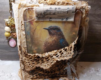 Puede incluir: Un diario hecho a mano con una ilustración de un pájaro. El diario está adornado con encaje, retazos de tela y cuentas. La portada presenta una pintura detallada de un pájaro con plumas marrones y azules. Tiene una estética vintage y rústica.