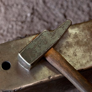 Pode incluir: Um close-up de um martelo de metal com um cabo de madeira descansando sobre uma bigorna. O martelo tem uma aparência gasta e envelhecida com uma pátina de ferrugem.