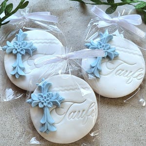 May include: Three white, round cookies with the word "Taufe" embossed on them, each topped with a light blue cross. The cookies are individually wrapped in clear cellophane and tied with white ribbons. Greenery is in the background.