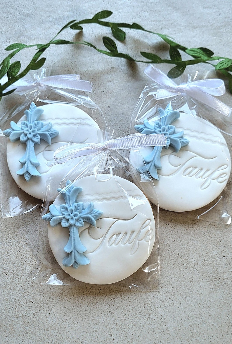 May include: Three white, round cookies with the word "Taufe" embossed on them, each topped with a light blue cross. The cookies are individually wrapped in clear cellophane and tied with white ribbons. Greenery is in the background.