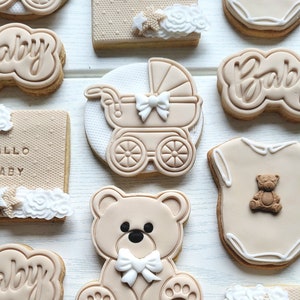 May include: Assortment of decorated sugar cookies with baby themes. Cookies feature a teddy bear, a baby carriage, a onesie, and the word "baby" in various styles. Some cookies have white icing details and star sprinkles.