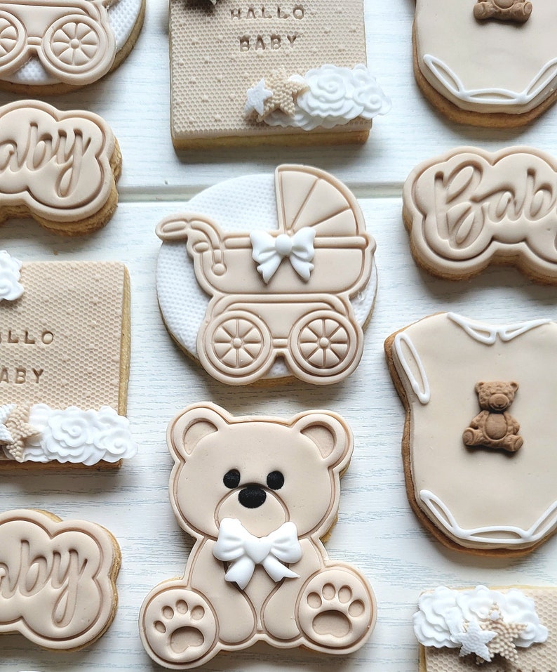 May include: Assortment of decorated sugar cookies with baby themes. Cookies feature a teddy bear, a baby carriage, a onesie, and the word "baby" in various styles. Some cookies have white icing details and star sprinkles.