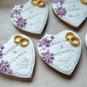 May include: Four white heart-shaped cookies decorated with purple and white flowers, gold rings, and the text "Marion & Michael".