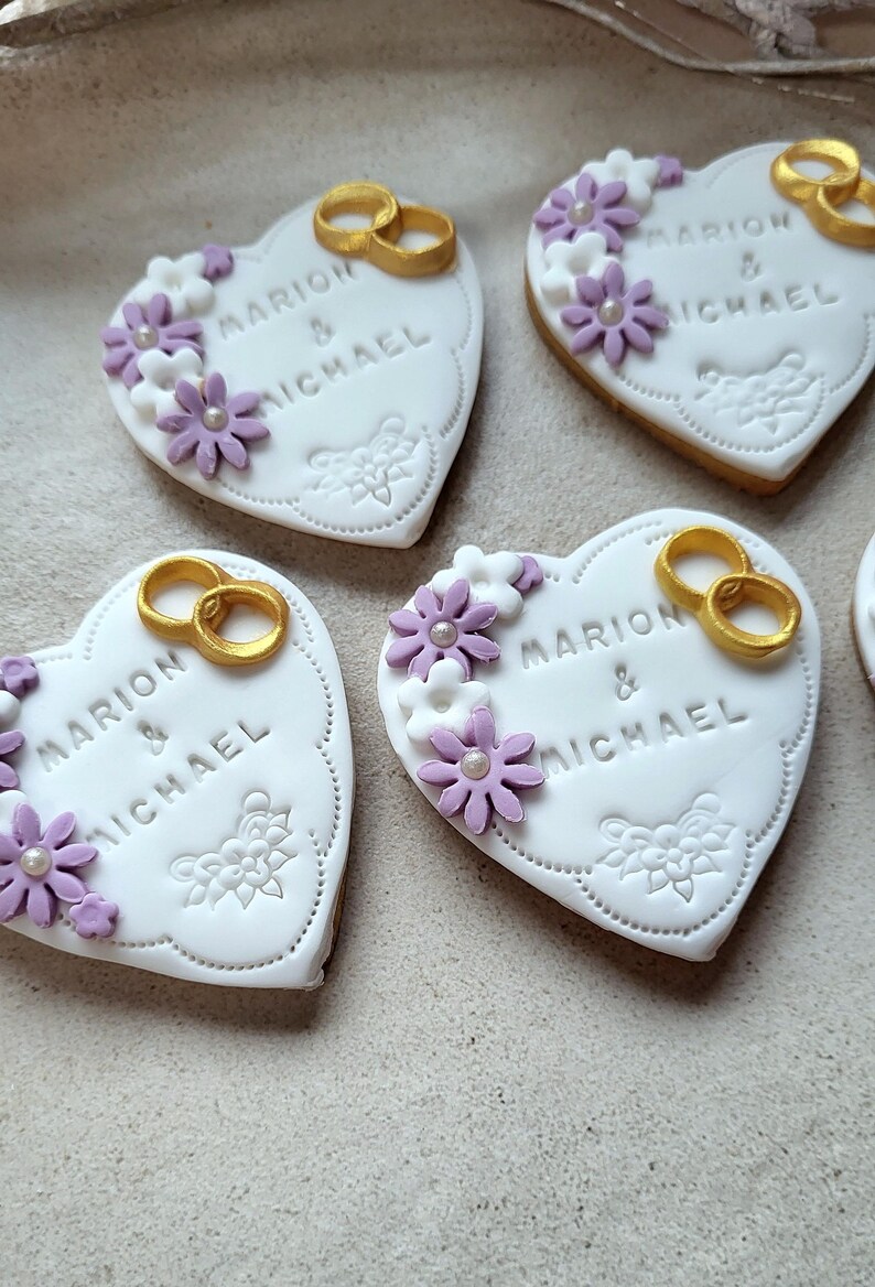 May include: Four white heart-shaped cookies decorated with purple and white flowers, gold rings, and the text "Marion & Michael".