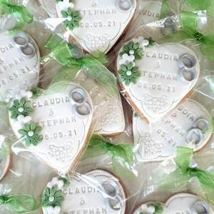 May include: Heart-shaped cookies decorated with white icing and green flowers. The cookies have the names "Claudia & Stephan" and the date "08.05.21" written on them. The cookies are packaged in clear plastic bags with green ribbon.