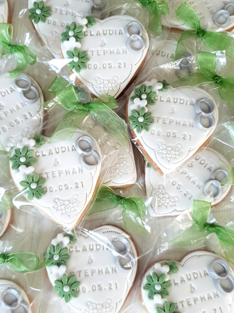 May include: Heart-shaped cookies decorated with white icing and green flowers. The cookies have the names "Claudia & Stephan" and the date "08.05.21" written on them. The cookies are packaged in clear plastic bags with green ribbon.